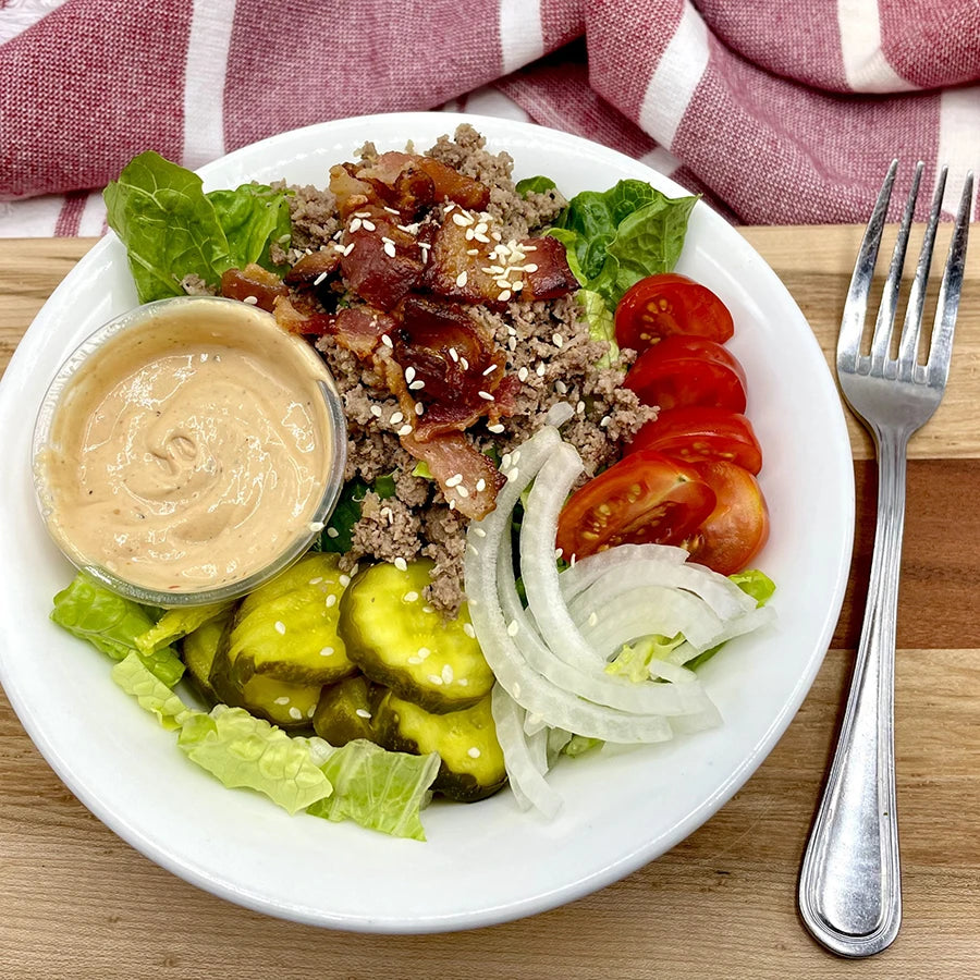 Plate with beef, bacon, tomatoes, onions, pickles, and a side of dressing.