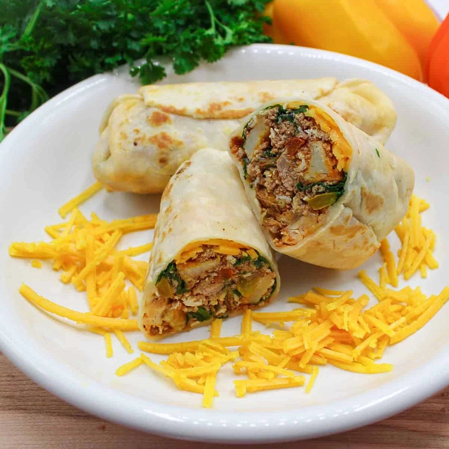 Two breakfast burritos on a plate with shredded cheese and vegetables in the background
