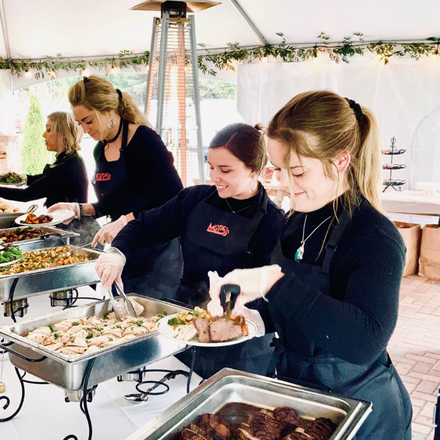 People serving food from a buffet table under a tented outdoor setting.