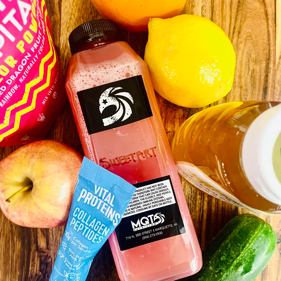 Bottle of pink juice with a label, surrounded by fruits and a protein powder packet.